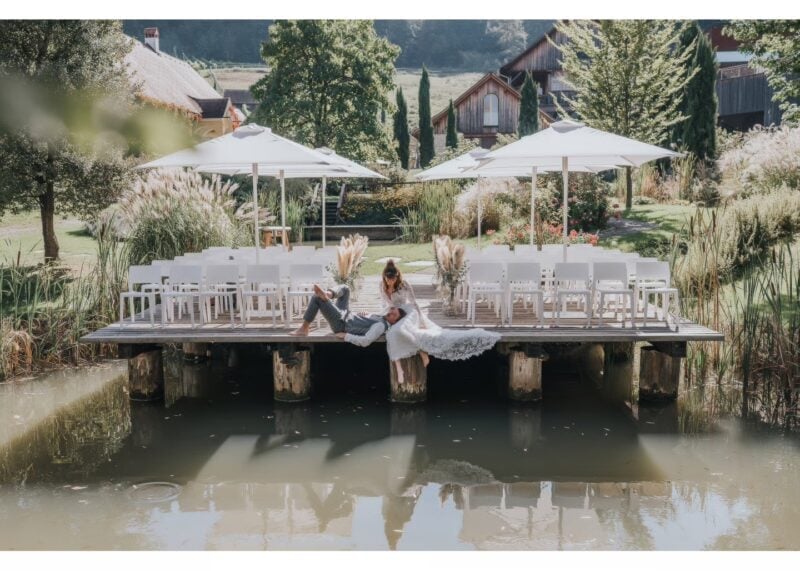 Kreatives Hochzeitsfoto: Brautpaar liegt entspannt auf einem Steg am Teich des Weinguts Georgiberg, dahinter die Hochzeitstafel