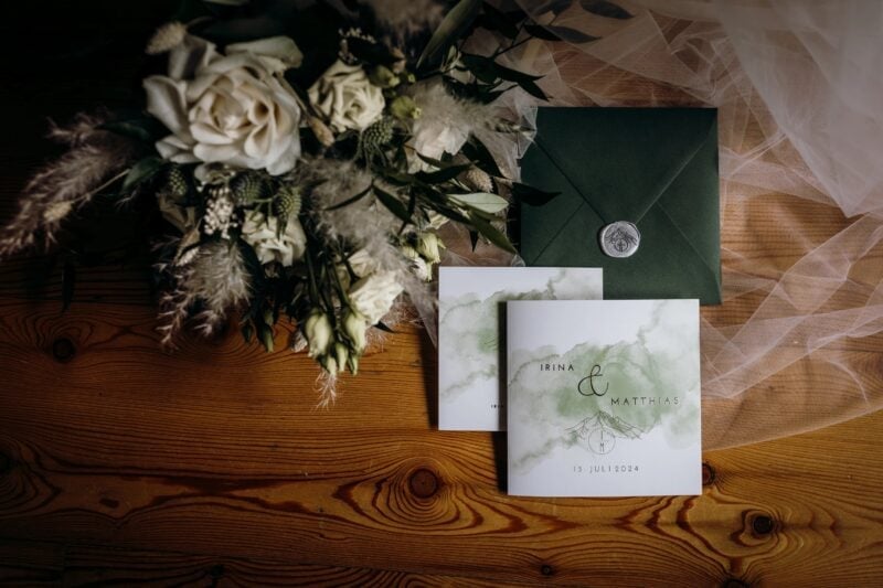 Hochzeitseinladung mit grünem Umschlag und weißem Brautstrauß auf Holztisch – Hochzeitspapeterie mit Zitat