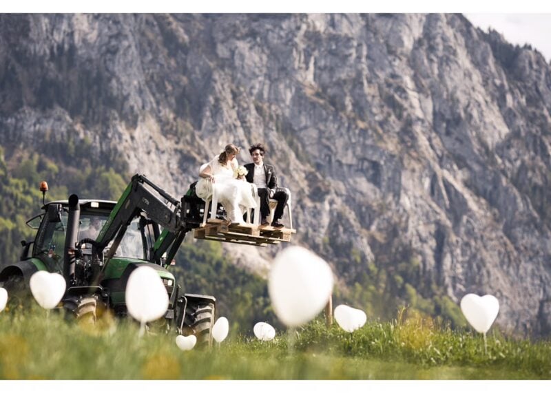 Brautpaar sitzt auf einem Traktor vor Bergkulisse, weiße Herzballons auf der Wiese – Hochzeit am Feldbauernhof