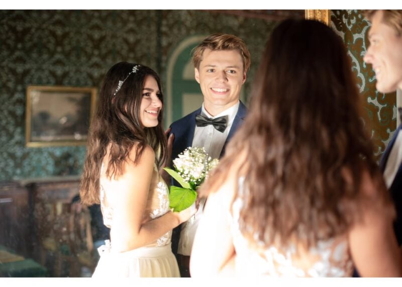 Braut mit Blumenstrauss und Bräutigam mit Gästen im historischen Salon von Schloss Reichenau