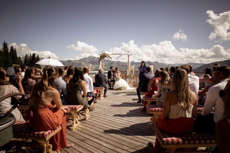 Hochzeitsgäste auf karierten Bänken verfolgen die freie Trauung auf der Terrasse des Madrisa-Hofs vor Bergkulisse