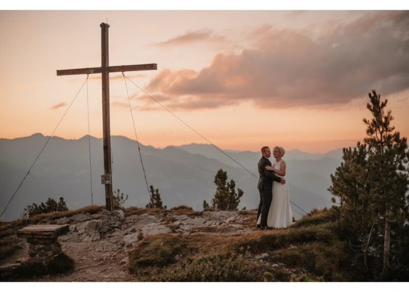 Brautpaar umarmt sich bei Sonnenuntergang am Gipfelkreuz – Berghochzeit beim Berghotel Gerlosstein
