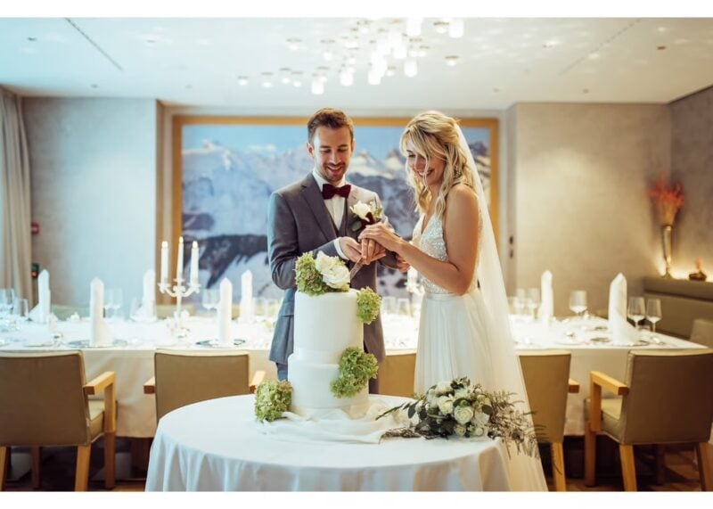Brautpaar schneidet die Hochzeitstorte im festlich gedeckten Saal des Kempinski Palace Engelberg an, dahinter die Gästetische