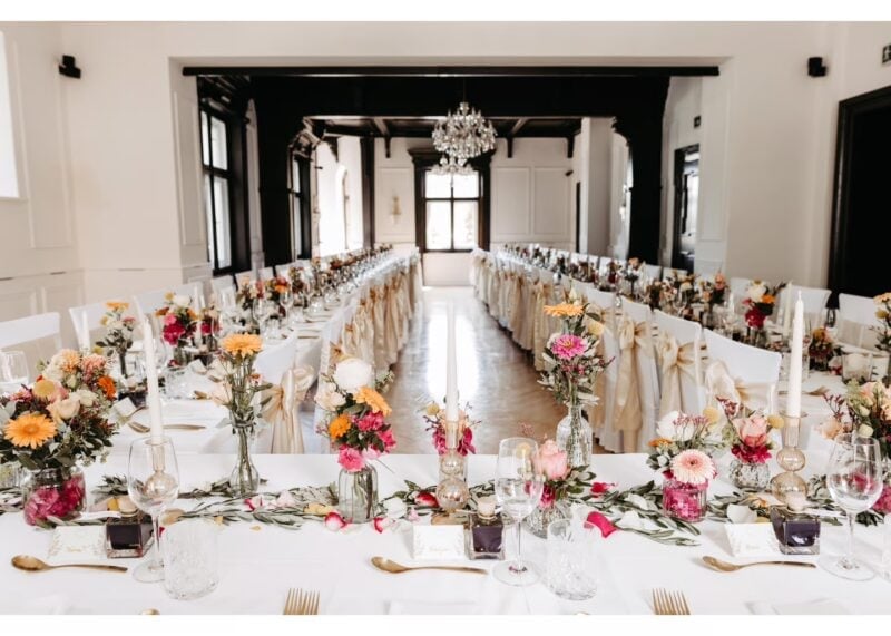 Lange Hochzeitstafeln mit bunten Blumen und Kerzen im hellen Festsaal des Georgi Schloss – Sitzordnung an langen Tafeln
