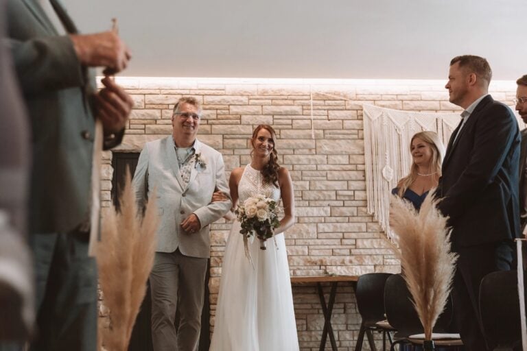 Vater begleitet lächelnde Braut beim Einzug, Location mit Steinwand und Trockenblumen