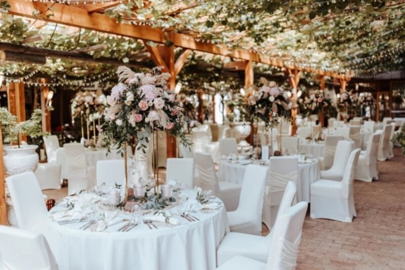 Runde Hochzeitstische mit Hussen und Rosen-Gestecken unter einer bewachsenen Pergola im Weingut Winzerhotel Kolleritsch