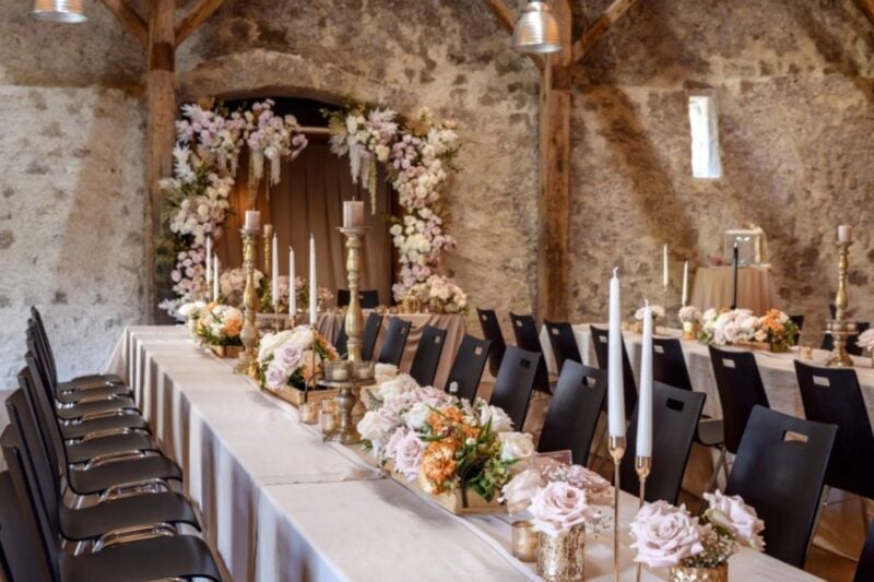 Lange Hochzeitstafel mit Kerzen und Rosen-Gestecken in der Trotte Villigen, dahinter ein Blumenbogen – Sitzordnung an einer Tafel