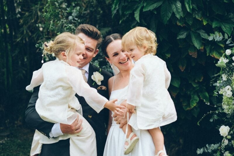 Lachendes Brautpaar hält zwei kleine Kinder in weissen Outfits auf dem Arm – Kinder bei der Sitzordnung der Hochzeit mitplanen