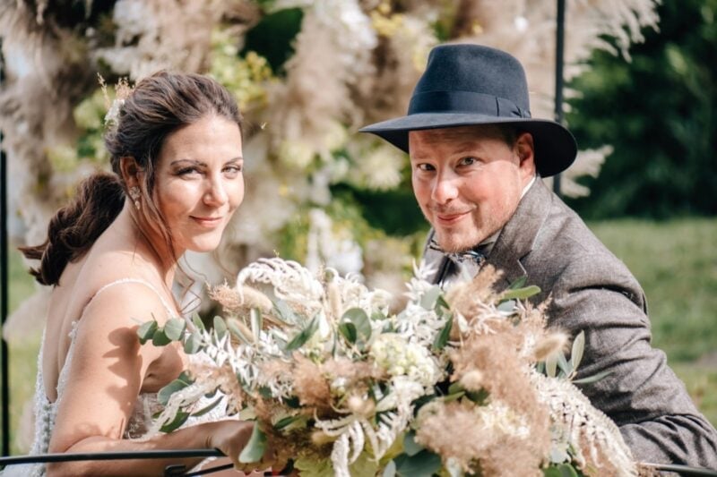Lächelndes Brautpaar mit Hut und üppigem Trockenblumen-Brautstrauss vor einer Blumenwand im Belvédère Strandhotel