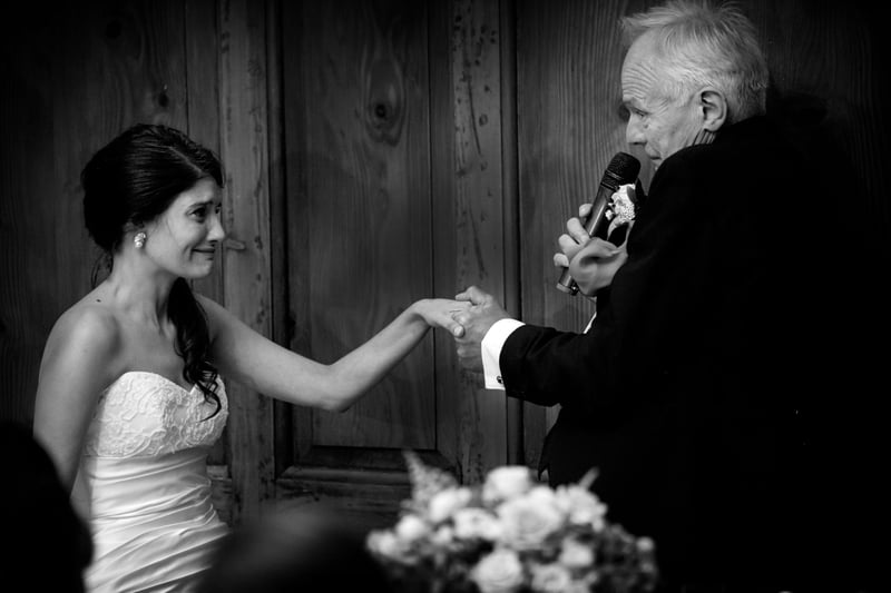 a father of the bride holds his daughter's hand as he gives his speech, both are emotional.