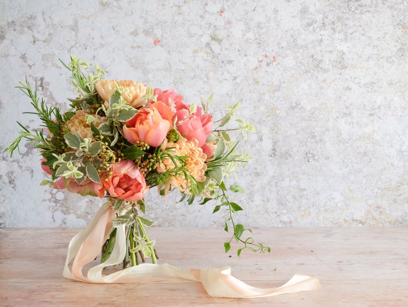 a pink, peach and green hand-tied bridal bouquet with trailing peach ribbon and foliage. Peonies are the main flower present.