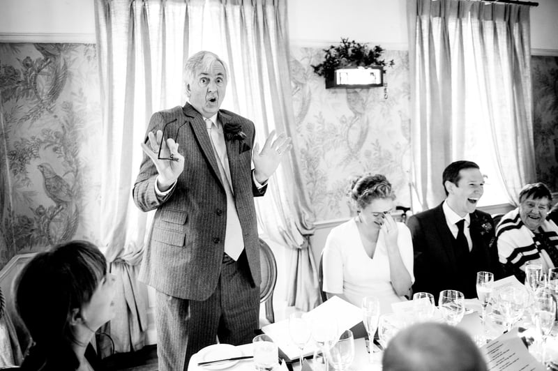 a father of the bride stands and gives a funny speech with hand gestures as the newlyweds laugh next to him.