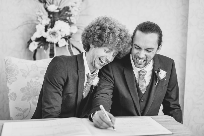 two grooms laughing as they sit and sign the register at their wedding