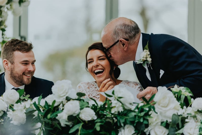 a father of the bride kisses his daughter on the cheek at the top table as she and her groom laugh.