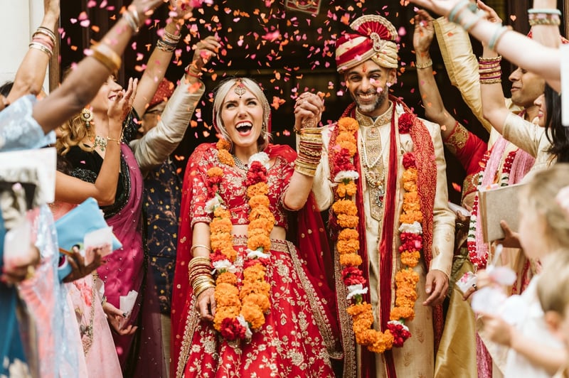 a bride and Indian groom leave their wedding ceremony in traditional Hindi dress and floral garlands as guests throw petal confetti