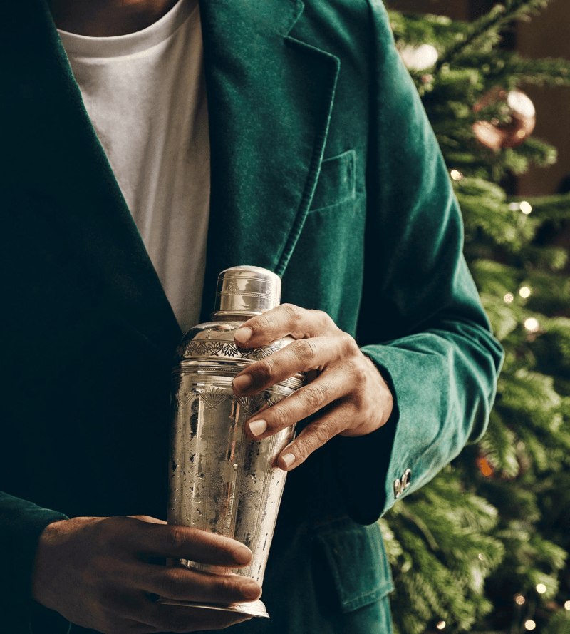 Man in a green velvet jacket holding an engraved silver cocktail shaker