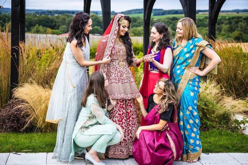 a hindu bride in traditional dress stands with her female guests who are all wearing colourful traditional Indian outfits.