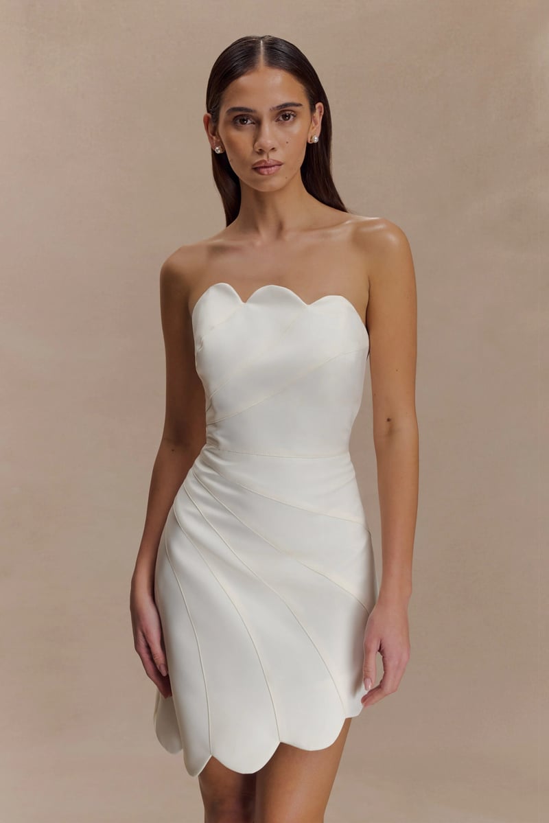 a model wears a white satin-style mini wedding dress with scalloped edging.