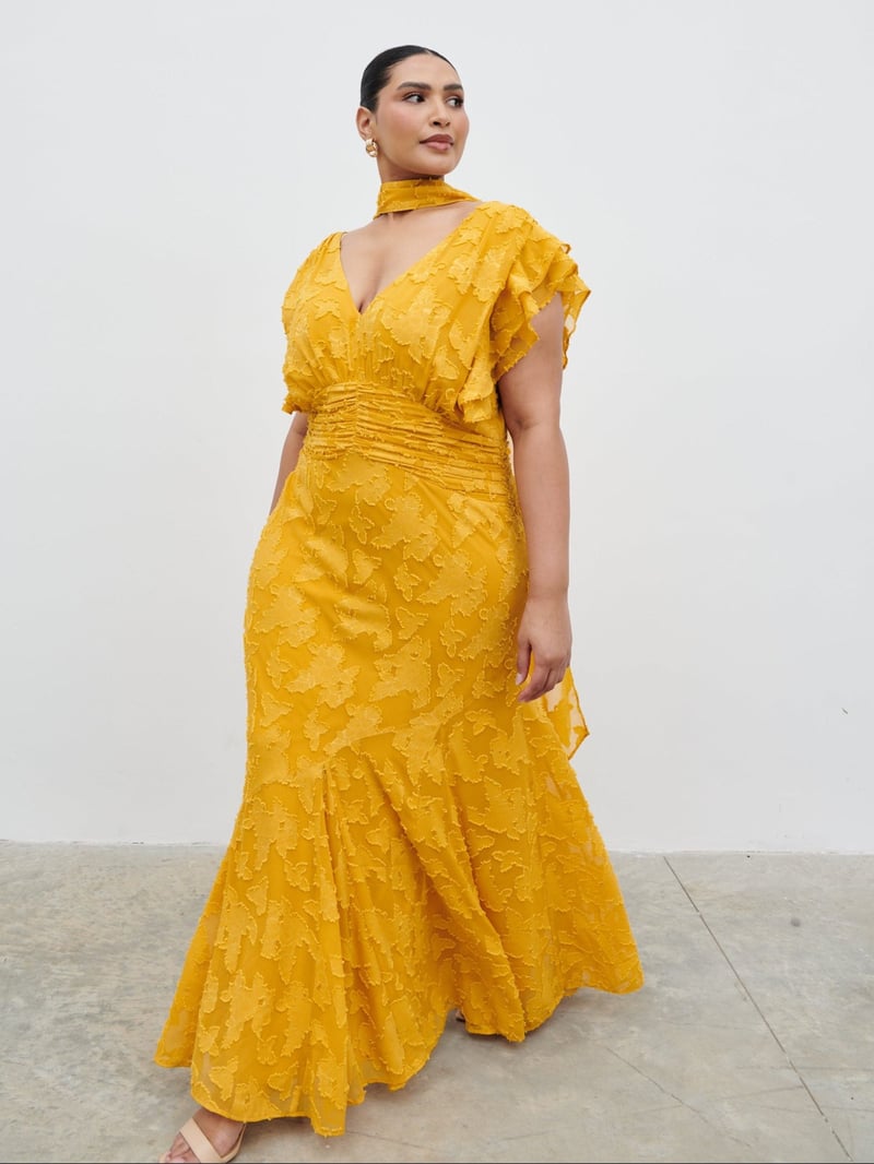 a plus size model wears a long ruffled maxi dress with scarf detail and short flutter sleeves in yellow ochre.