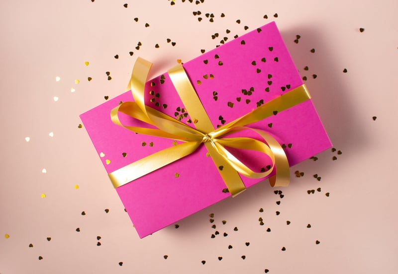 rectangular gift wrapped in pink wrapping paper and a gold ribbon bow, covered in pink heart confetti
