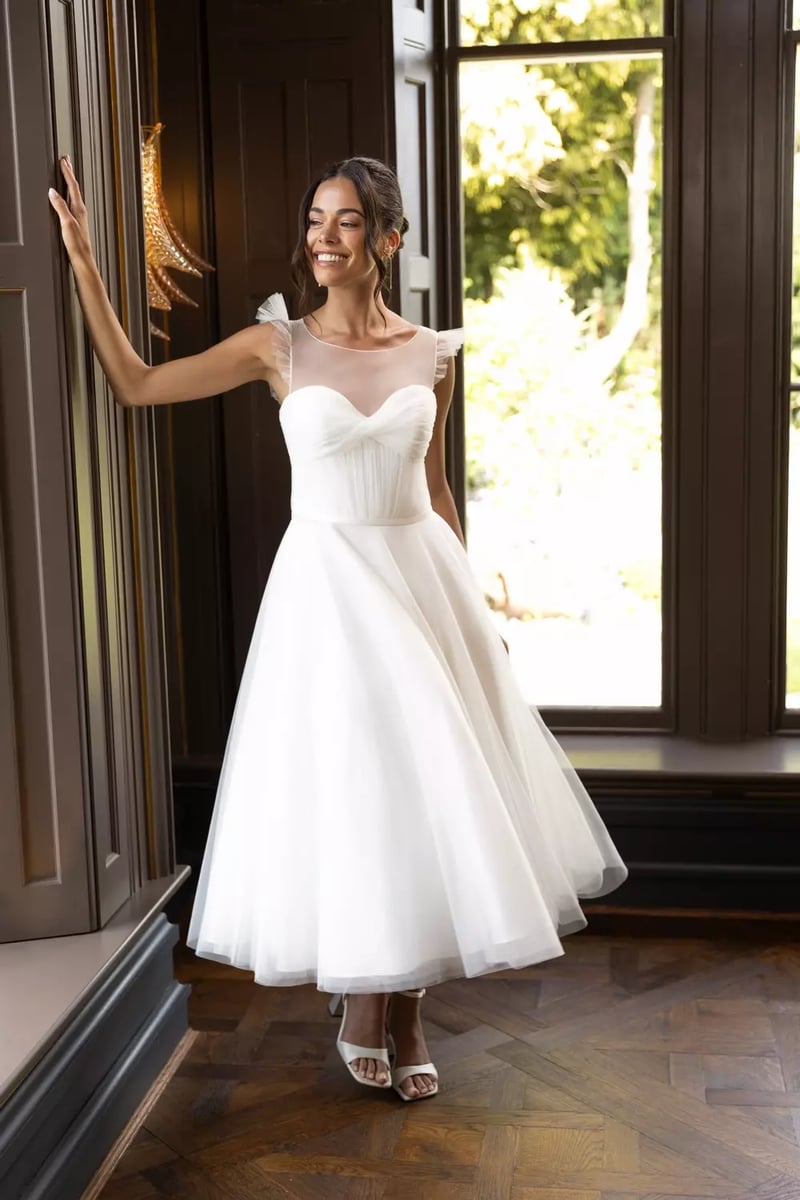 a model looks out of the window and smiles. She wears a tea-length wedding dress with full skirt and chiffon overlay with illusion neckline on the bodice