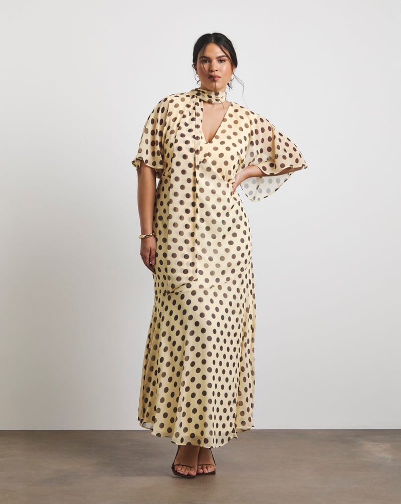 a plus size model wears a butter yellow and brown polka dot maxi dress with scarf detail.