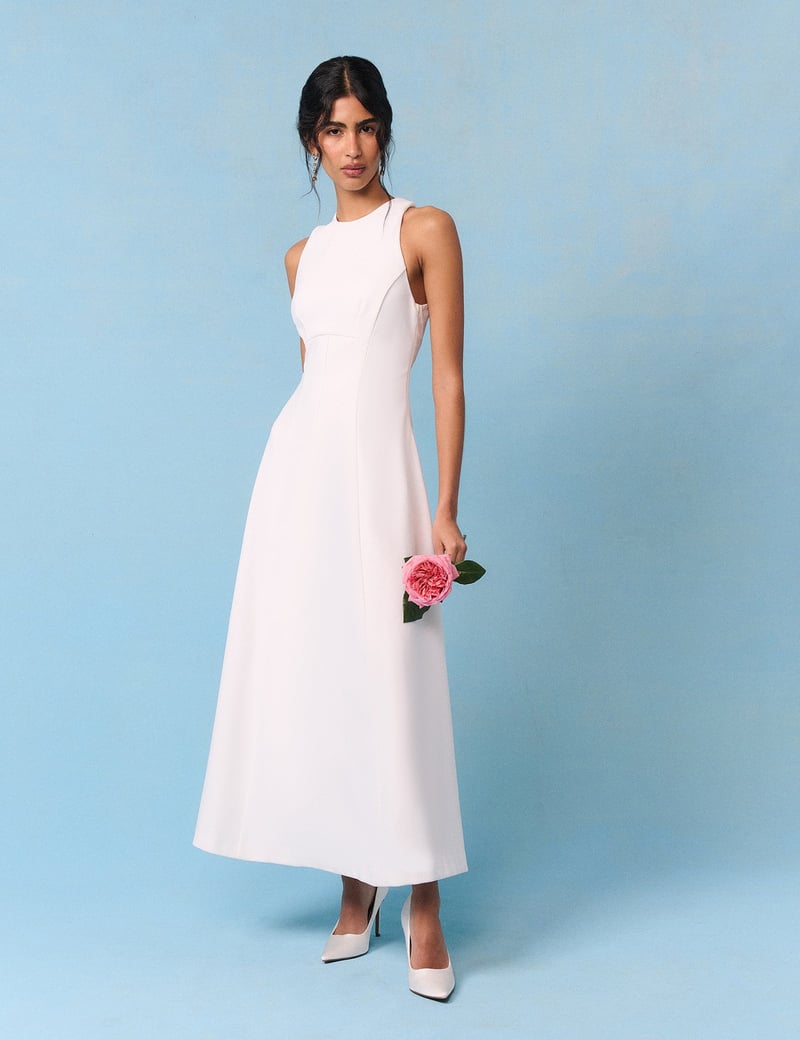 a model wears a white crepe fabric tea length wedding dress with high neckline. She holds a small posy of flowers.