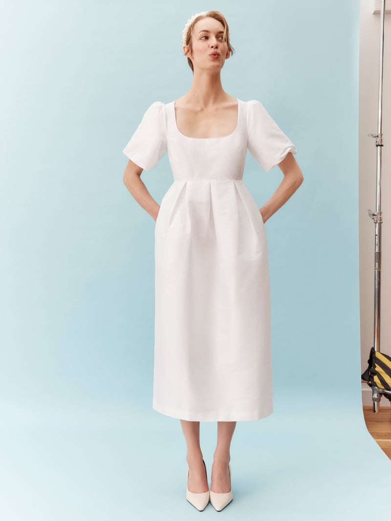 a model stands with her hands in her pockets wearing a white midi wedding dress with short puff sleeves and square neckline.