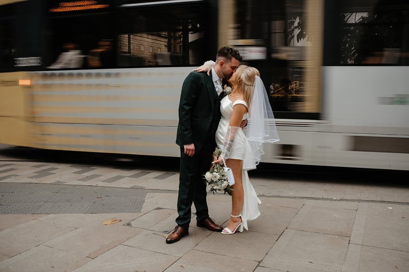 a bride wearing a short wedding dress kissing her groom as a bus drives past in the background.
