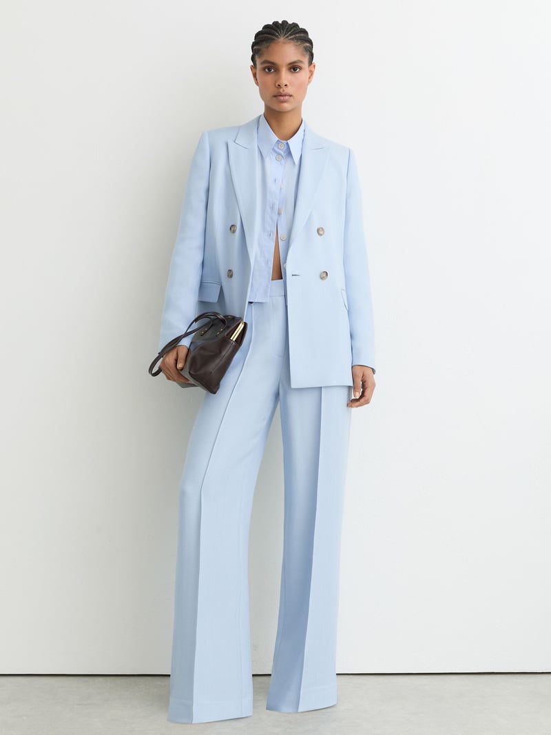a model wears a pale blue double breasted trouser suit and holds a black clutch bag.