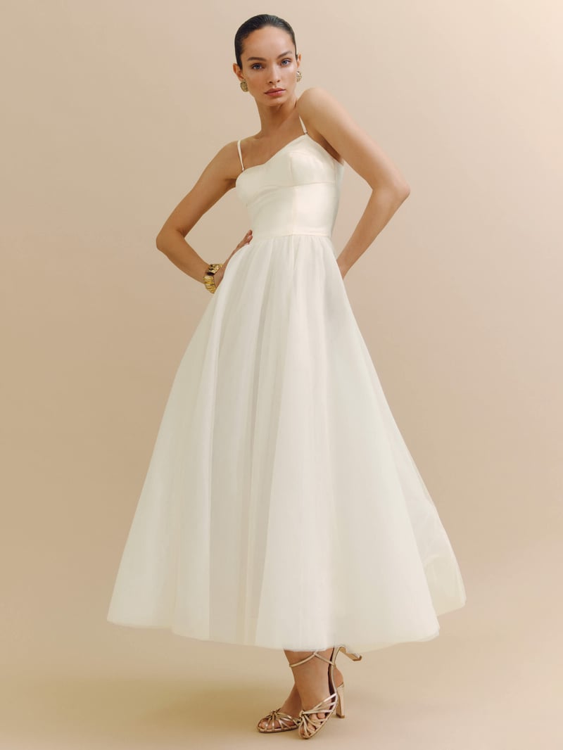 a model stands with her hands on her hips wearing a full skirted tulle tea length wedding dress with satin strappy bodice.