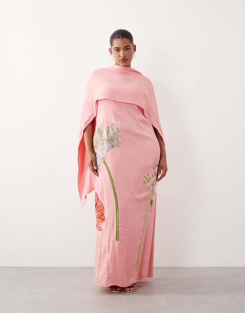 a plus size model wears a pale pink satin maxi dress with high neck cape detail and embroidered florals.