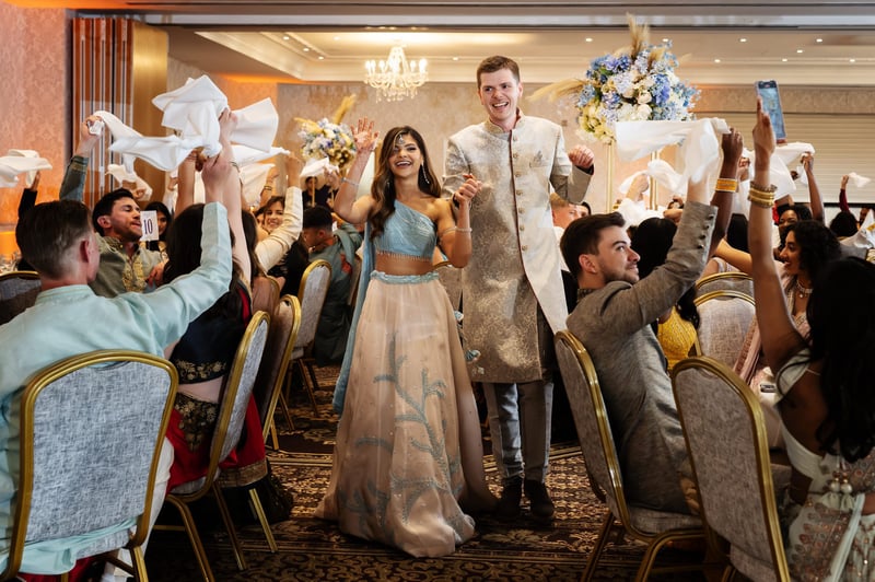a hindu bride and her groom walk hand in hand through their wedding breakfast as guests wave their napkins in the air.