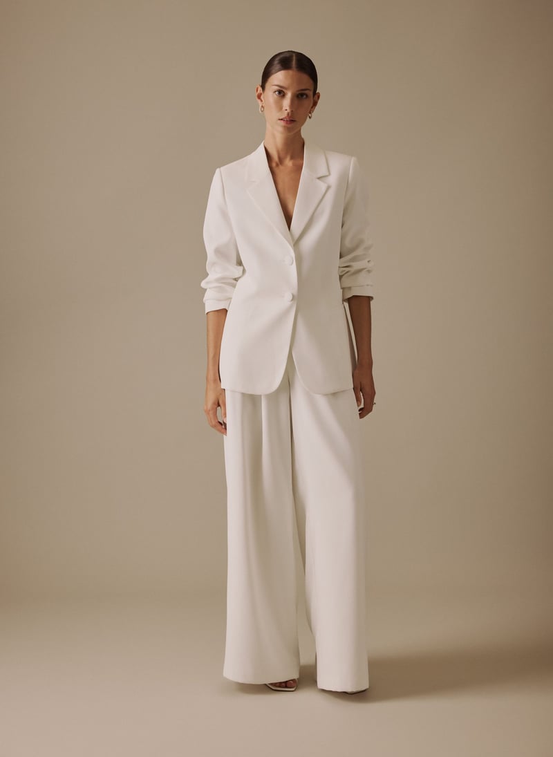 a model wears an ivory coloured two-piece wedding suit with single breasted blazer and wide-leg trousers.