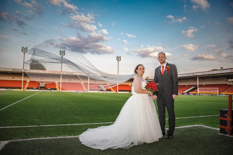 A bride and groom standing on the pitch at Walsall Football Club's stadium, with the stadium stands and floodlights visible behind them