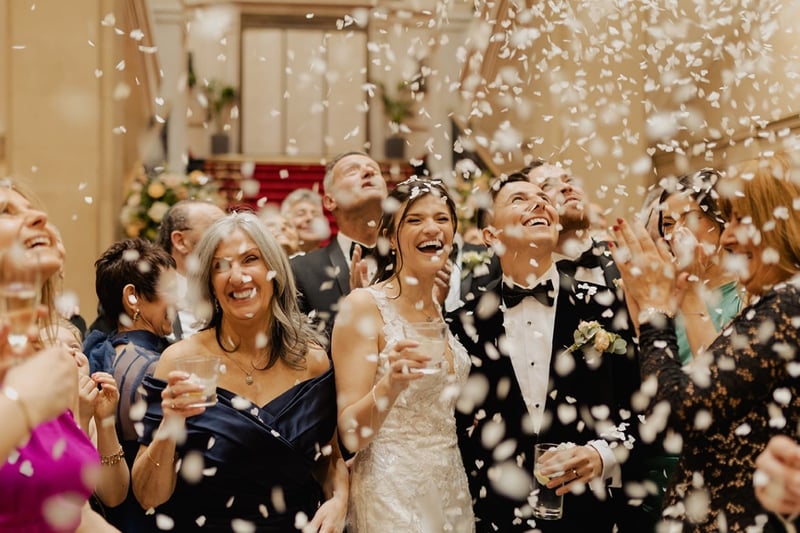 a bride and groom smile as they are surrounded by their guests and confetti falls from the sky.