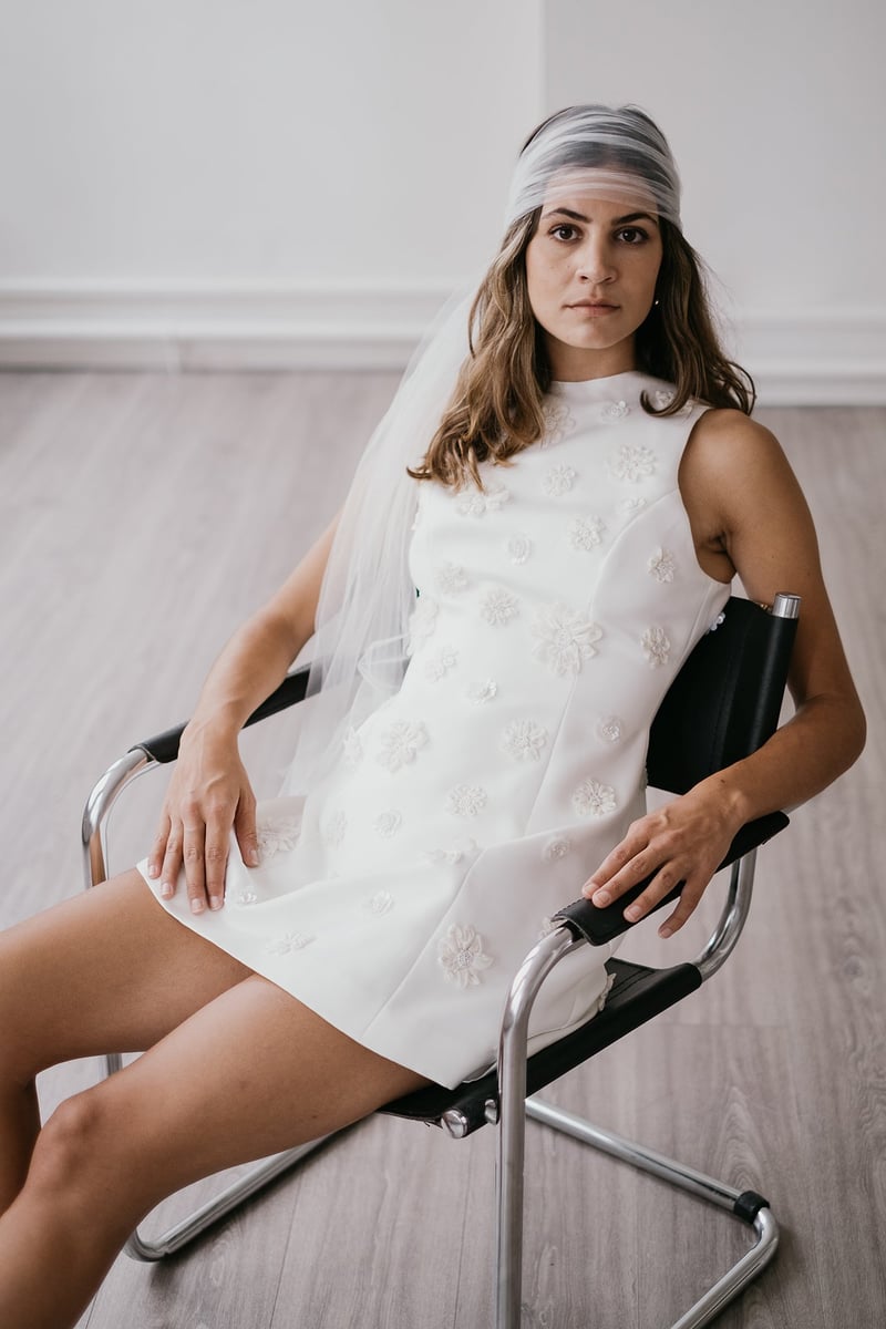 a model sits in a chair wearing a short, sleeveless embellished wedding dress with veil.