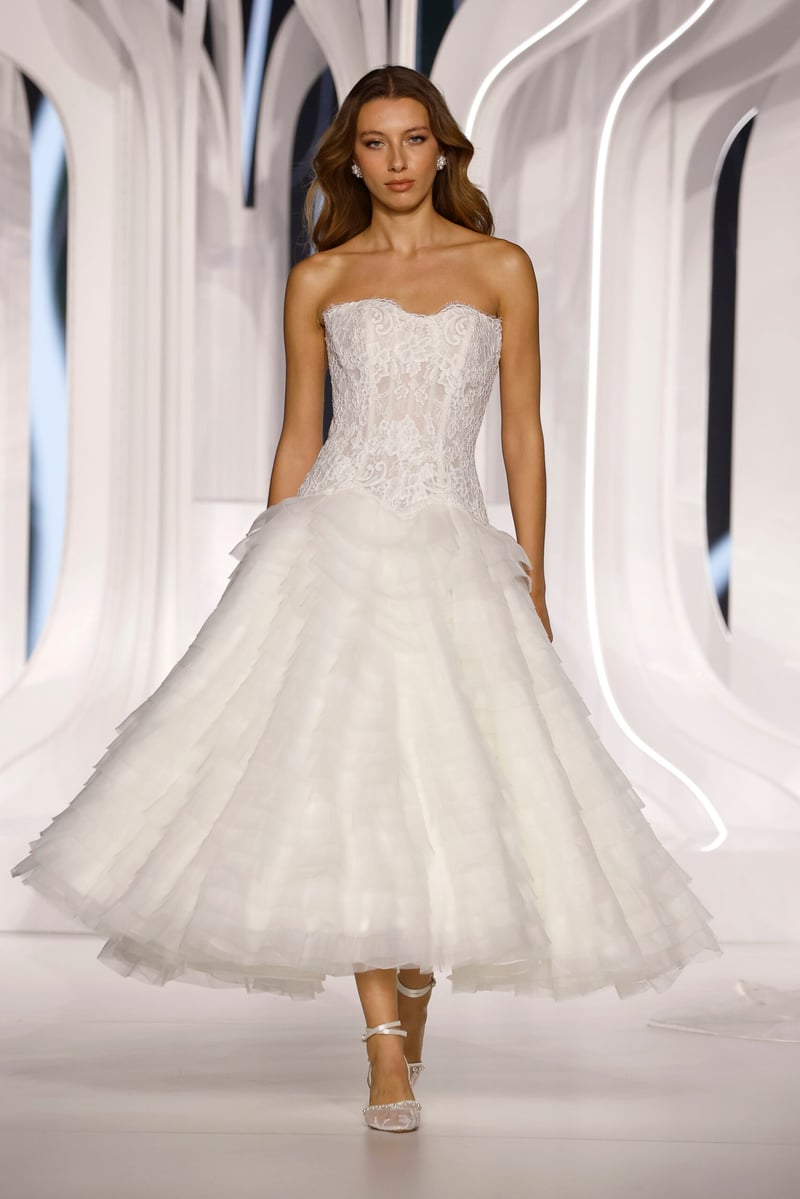 a model walks down the runway wearing a strapless tea length wedding dress with full tulle skirt and lace corset.