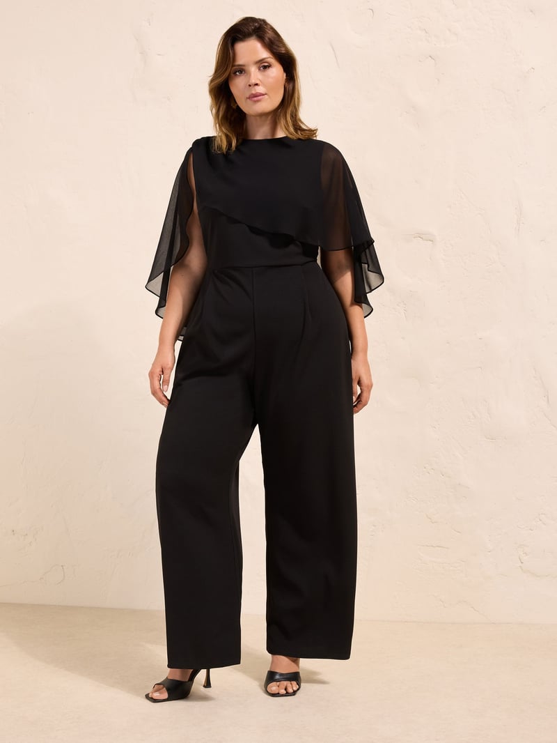 a plus size model wears a black tailored jumpsuit with chiffon cape sleeves.