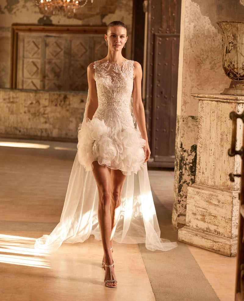 a model wearing a short embellished wedding dress with ruffled organza trim and a detachable cape.