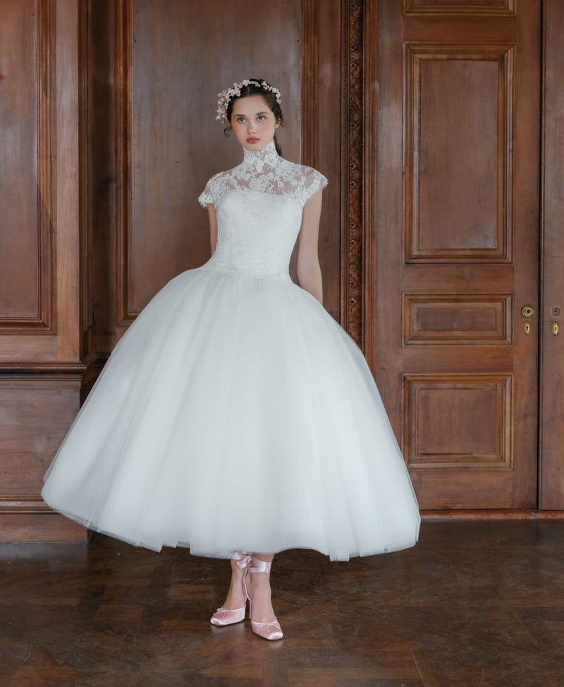 a model wears a 1950s-style tea length wedding dress with tulle skirt and high neck lace bolero.