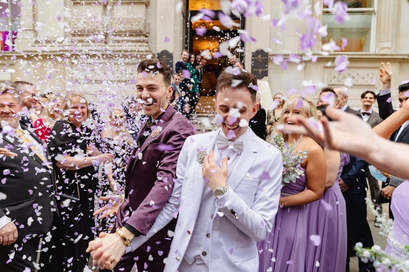 two grooms at an LGBTQ+ wedding in purple suits walk hand in hand outside their wedding venue as guests throw confetti.
