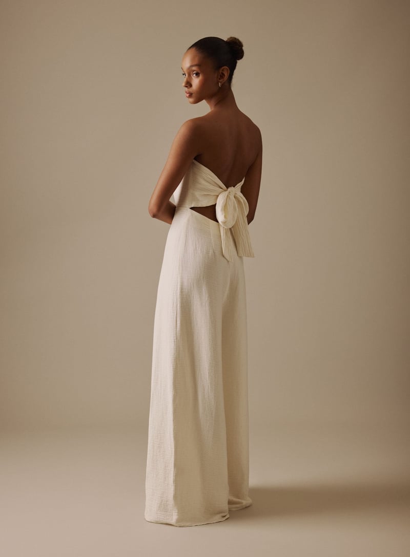 a model stands with her back to the camera wearing a cream wide leg jumpsuit with open tie-back detail.