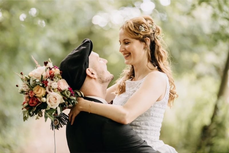 Braut mit lockerer Half-Up-Brautfrisur und floralem Haarschmuck lächelt ihren Bräutigam bei einem emotionalen Hochzeitsshooting im Grünen an.