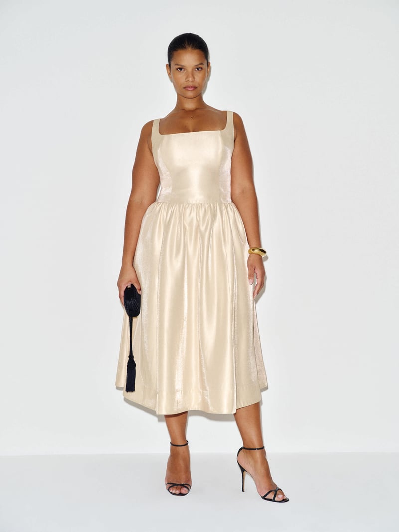 a model wears a silk champagne-coloured midi wedding dress with square neckline and holds a black clutch bag.