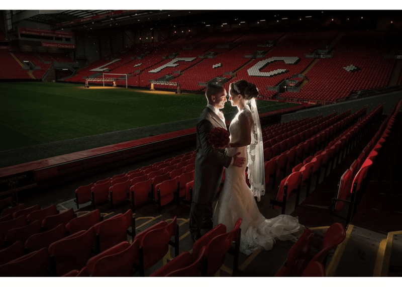 A bride and groom standing in the Anfield stadium stands, with the LFC lettering and pitch visible behind them