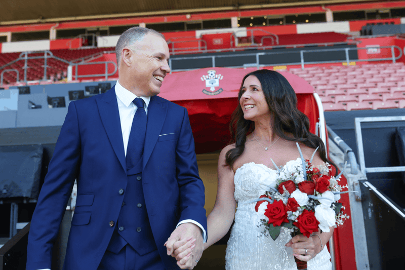 A smiling bride and groom in front of the stands at Southampton Football Club's St Mary's Stadium, with the bride holding a red and white bouquet