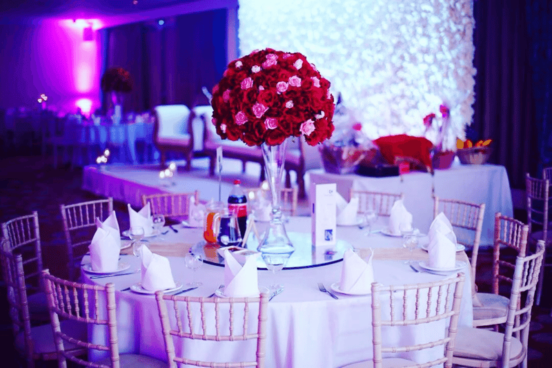 A wedding reception at Charlton Athletic Football Club's The Valley, with gold chiavari chairs and tall red rose centrepieces under purple mood lighting