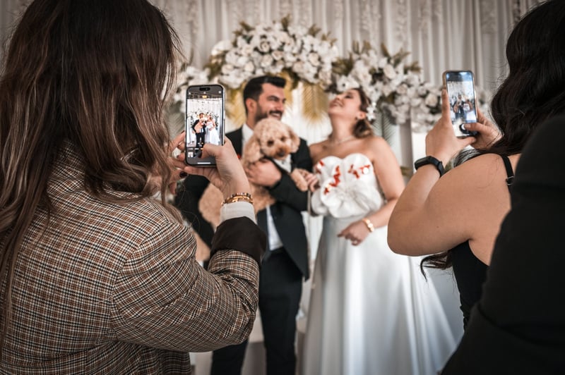 a bride and groom smile as they pose with their dog and guests take photos of them.