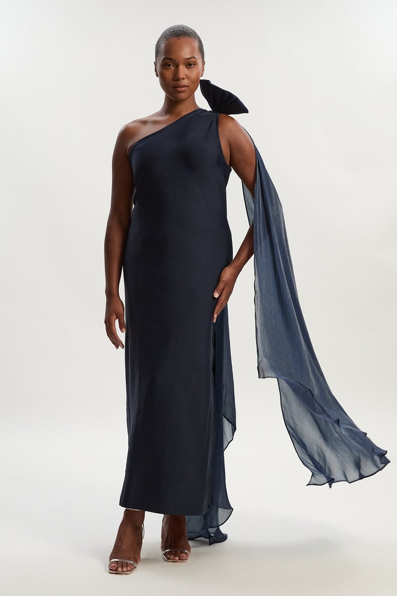 a curve model wears a navy blue knit-style bodycon maxi dress with bow and cape detail at the back.